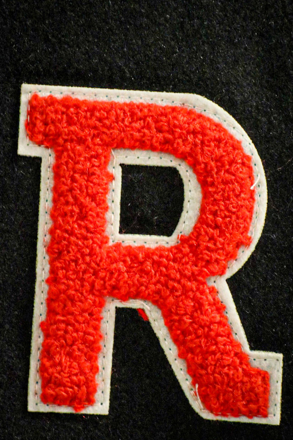 Close-up of the red 'R' chenille patch on the left chest of the black RMIT varsity jacket, featuring white trim around the letter, available at the RMIT Store.